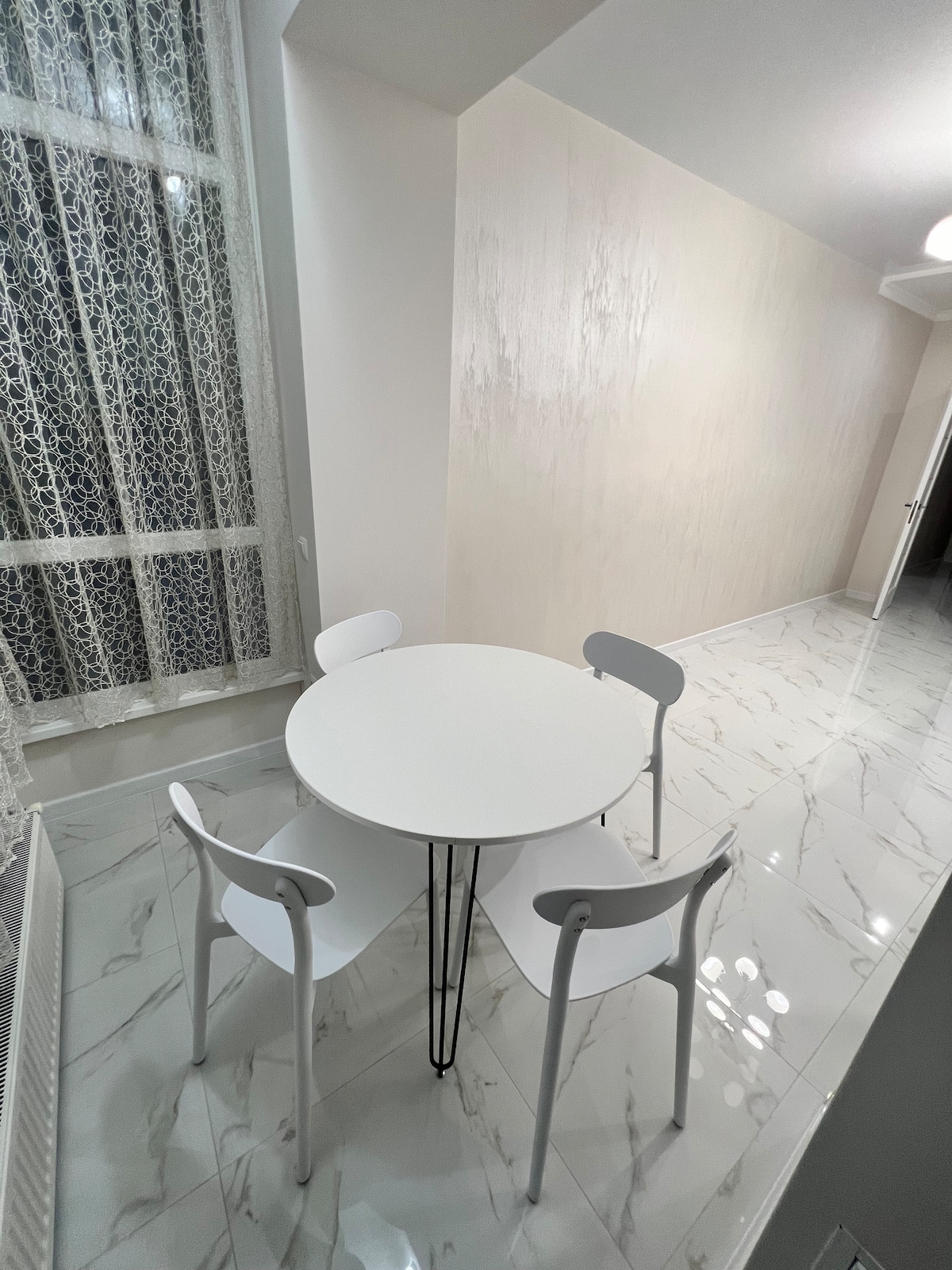 A dining area features a round table accompanied by three sleek white chairs. The floor is adorned with glossy tiles reflecting the light, and a window adorned with delicate lace curtains allows natural light to brighten the space.