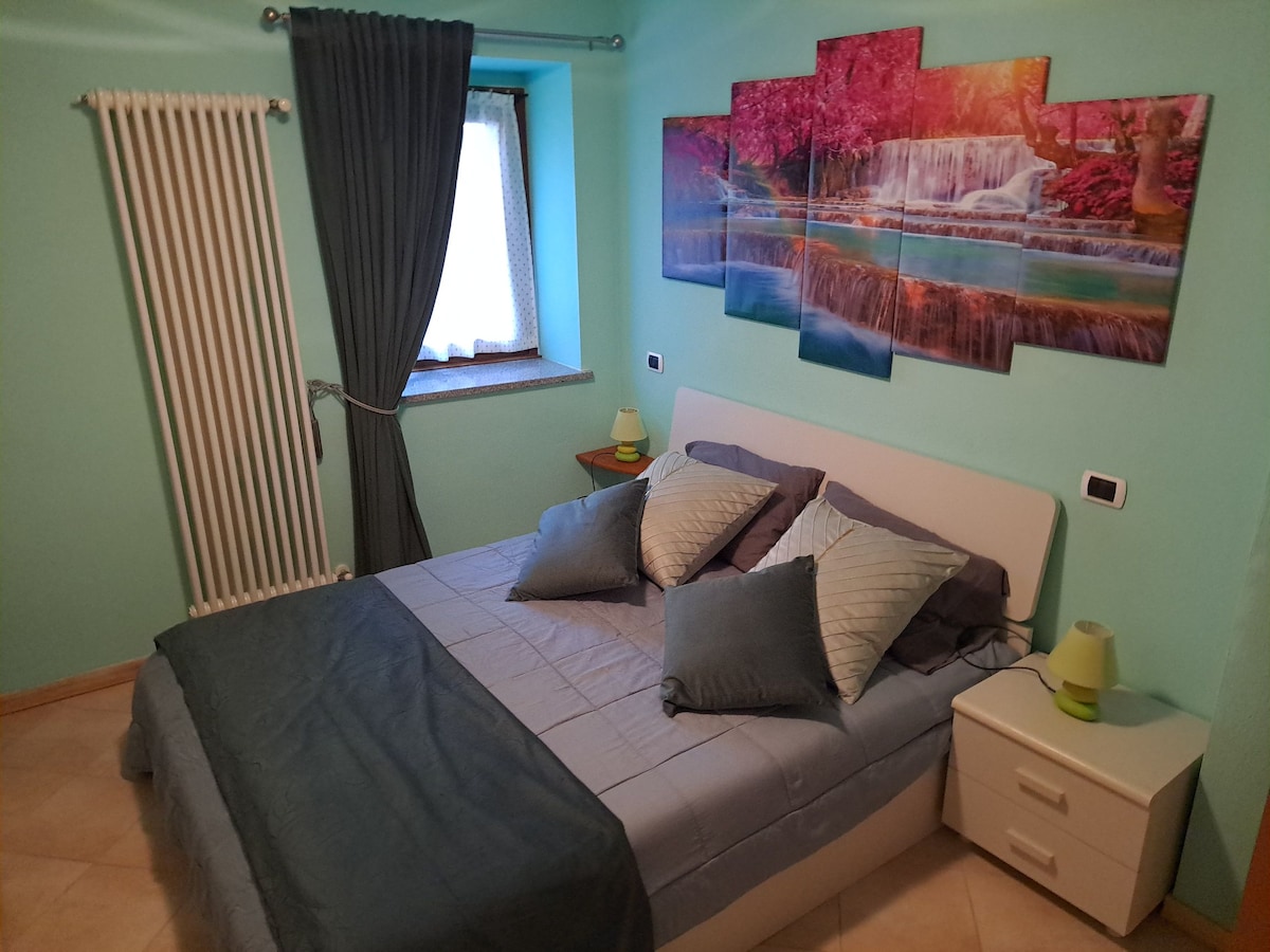 A cozy bedroom is presented with a spacious bed topped with decorative pillows. A piece of wall art depicting waterfalls and vibrant foliage enhances the atmosphere. A window allows natural light, complemented by dark curtains. A bedside table is situated next to the bed.