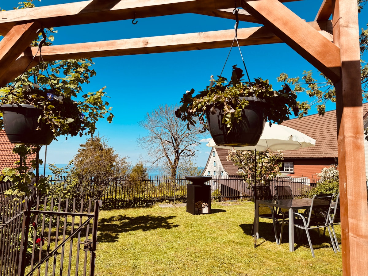 A private garden area is presented, featuring a well-maintained lawn bordered by a wooden fence. Hanging planters add greenery above a small outdoor dining set, consisting of a table and two chairs. An umbrella provides shade, with a pleasant view of the lake visible beyond.