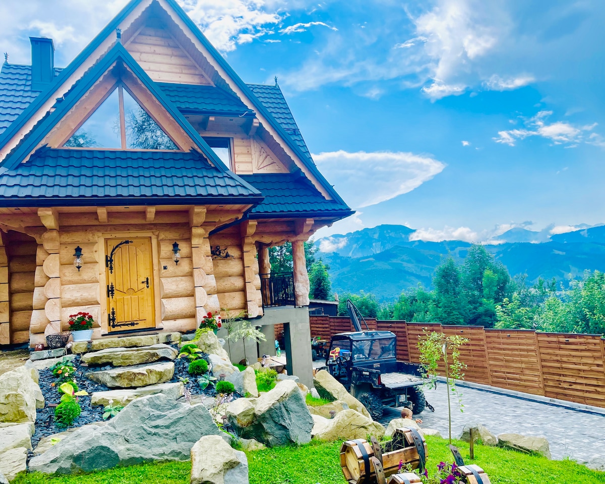 The exterior of the villa features a charming wooden structure complemented by landscaped greenery. A stone pathway leads to the entrance, which is adorned with a large yellow door. The backdrop showcases the mountains under a blue sky with scattered clouds.