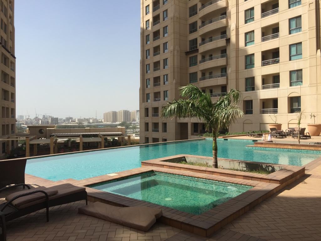 An outdoor pool area is depicted, featuring a large swimming pool with crystal-clear water, surrounded by a tiled deck. A palm tree is positioned near the edge of the pool, and a smaller hot tub is visible adjacent to the pool. The backdrop showcases modern buildings and a clear sky.