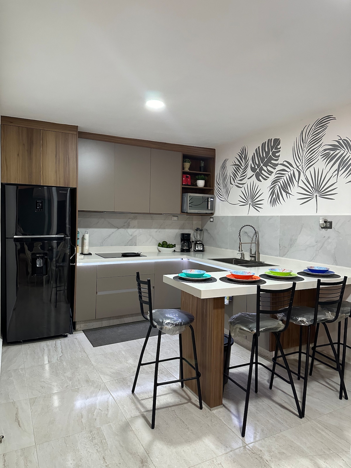 A modern kitchen features sleek cabinetry with a combination of wood and neutral tones. A central island with seating for four is set with colorful plates. Stainless steel appliances, including a refrigerator and coffee maker, are visible against a backdrop of elegant marble countertops.