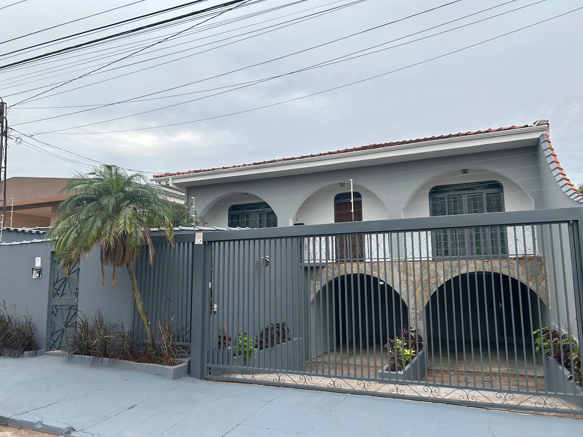The exterior view of the property features a two-story structure with a gated entrance. Archways create a welcoming entry, surrounded by a landscaped garden with low-growing plants. A palm tree adds a tropical touch to the setting, while the building is painted in a subtle grey hue.
