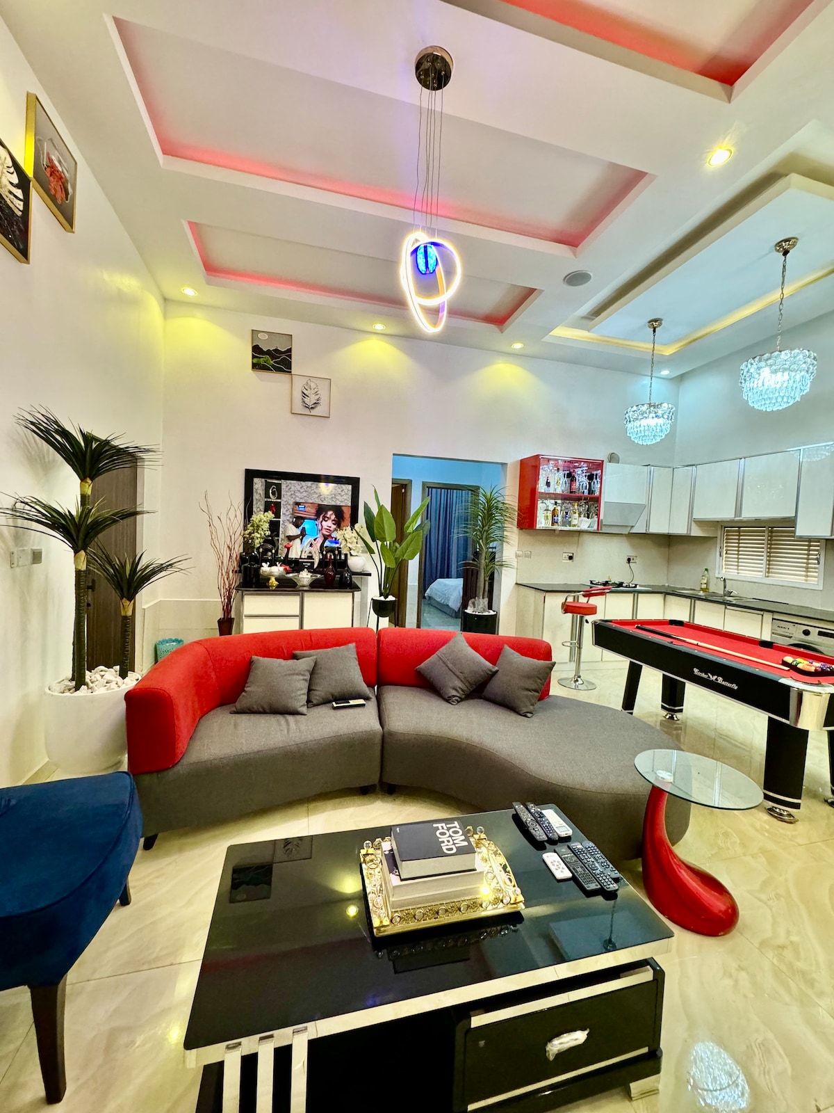 A modern living area features a curved gray sofa with red accents, arranged around a sleek glass coffee table. The space is enhanced by decorative plants and unique light fixtures. A billiard table is positioned in the background, complemented by stylish cabinetry in the kitchen area.