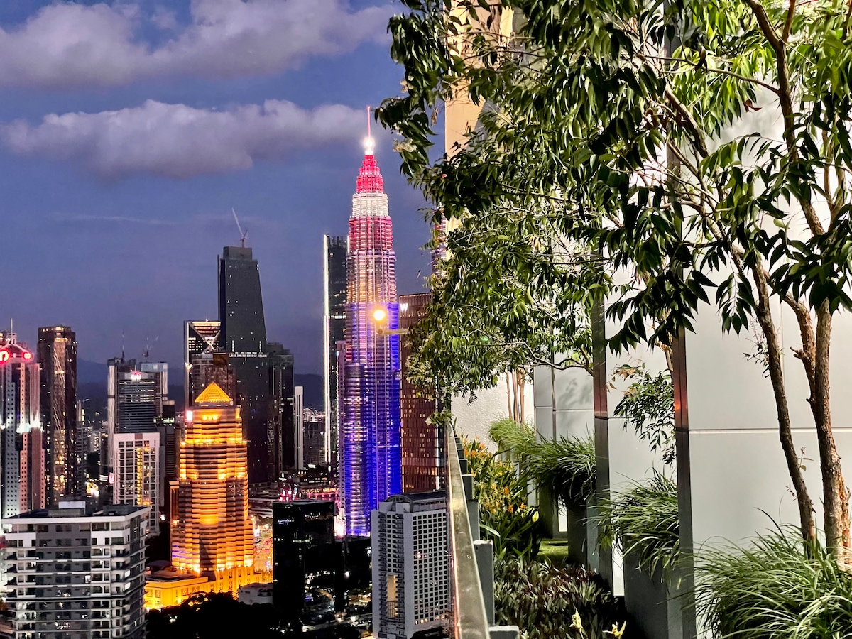 A panoramic view of Kuala Lumpur's skyline showcases vibrant lights from the city, including the iconic Petronas Twin Towers. Lush greenery is visible along the balcony edge, enhancing the modern architectural aesthetic of the apartment.