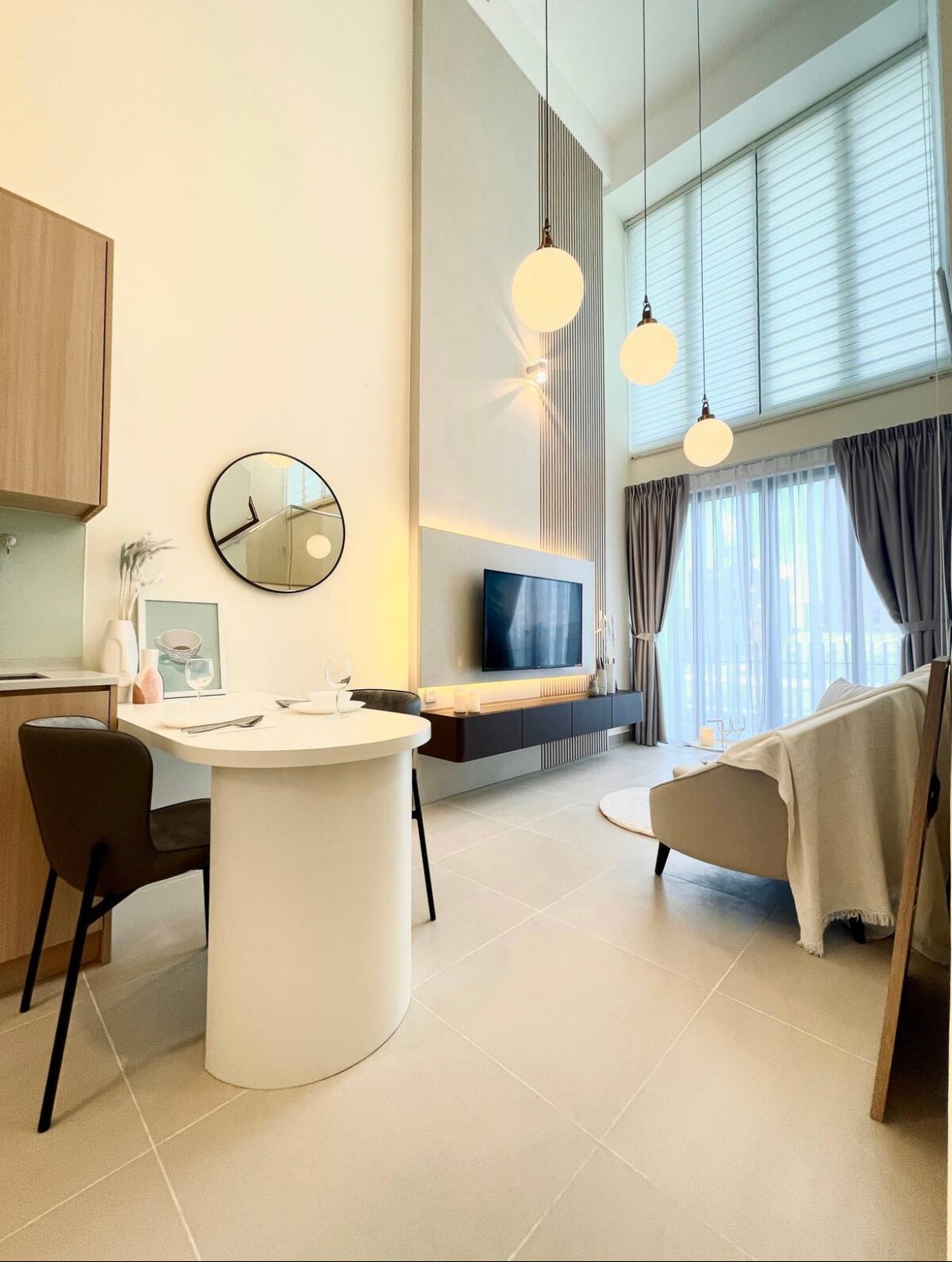 The dining area features a round table with two black chairs, complemented by a wall-mounted TV and stylish cabinetry. Natural light filters through large windows with sheer curtains, while pendant lights hang elegantly from the ceiling, creating a serene atmosphere.
