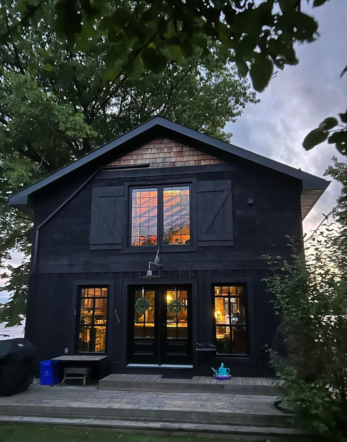 The exterior of a rustic cabin is showcased, featuring dark wood siding and large glass windows that reflect the sky at dusk. Two sets of double doors are centered at the bottom, framed by greenery, creating a warm and welcoming entrance.