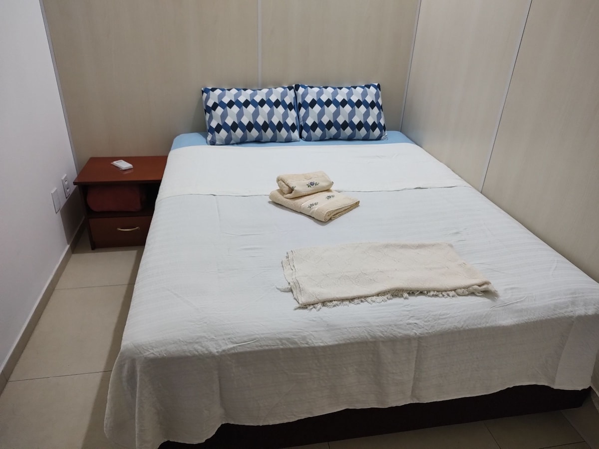 A neatly made bed is centered in the room, covered with a white blanket. Two decorative pillows with a geometric pattern rest against the headboard. A small nightstand is positioned beside the bed, showcasing additional linens on top.