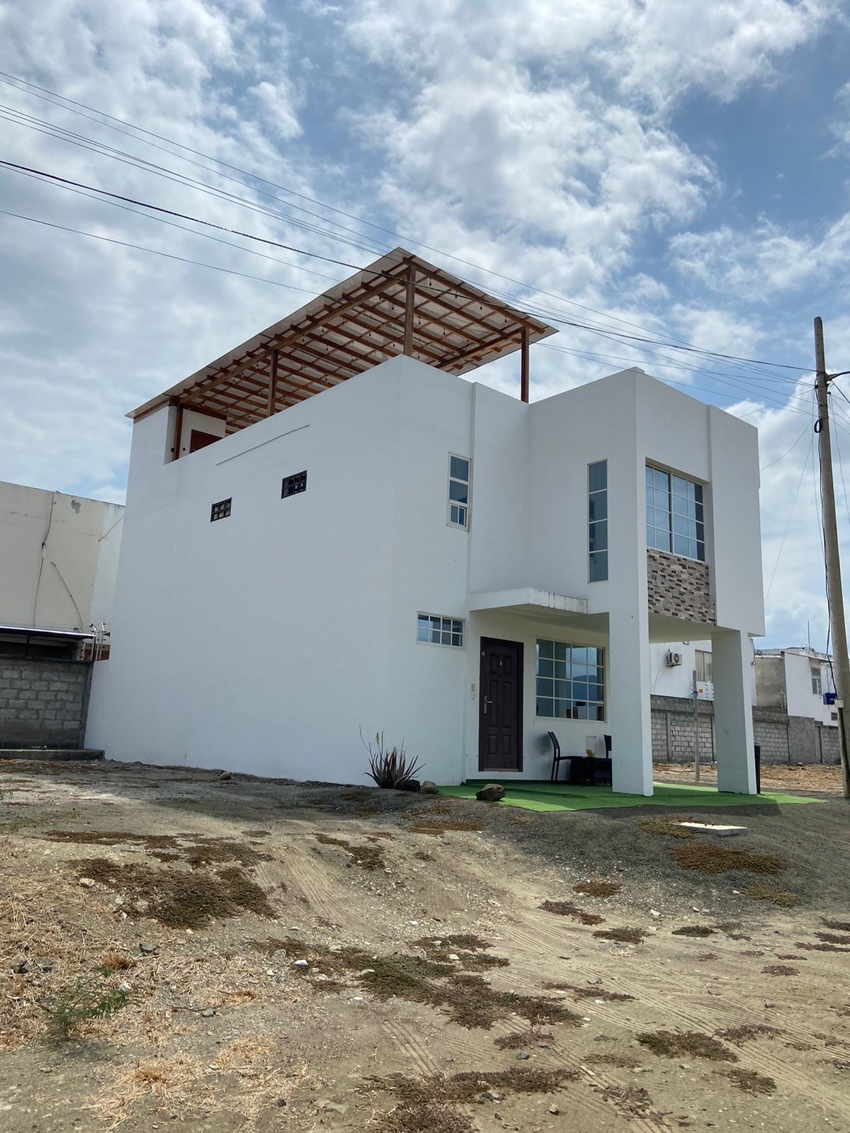 A modern, two-story house is depicted, featuring a clean white exterior with large windows. A flat roof structure provides an open area above. The surrounding landscape includes a dirt pathway and sparse vegetation, emphasizing the property's spaciousness.