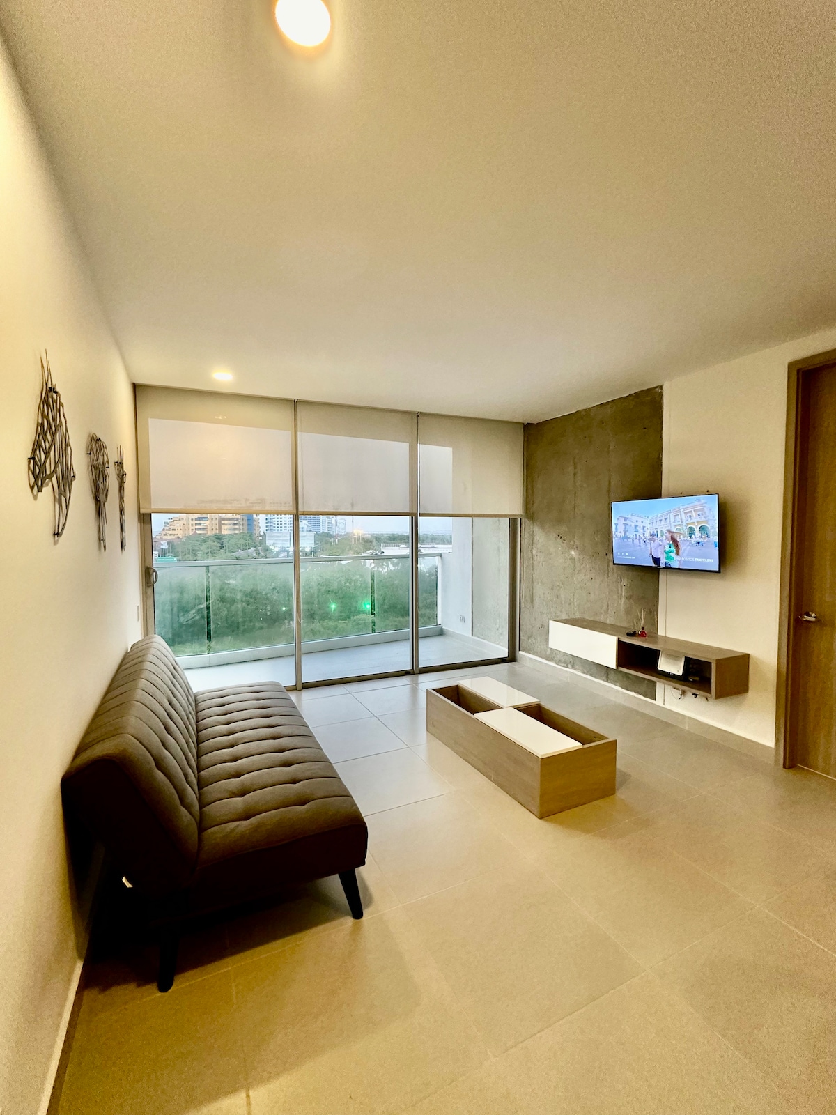 A modern living area features a comfortable sofa and a minimalist coffee table, positioned in front of a wall-mounted television. Large glass doors provide access to a private terrace, allowing natural light to fill the space while offering views of the surrounding area.