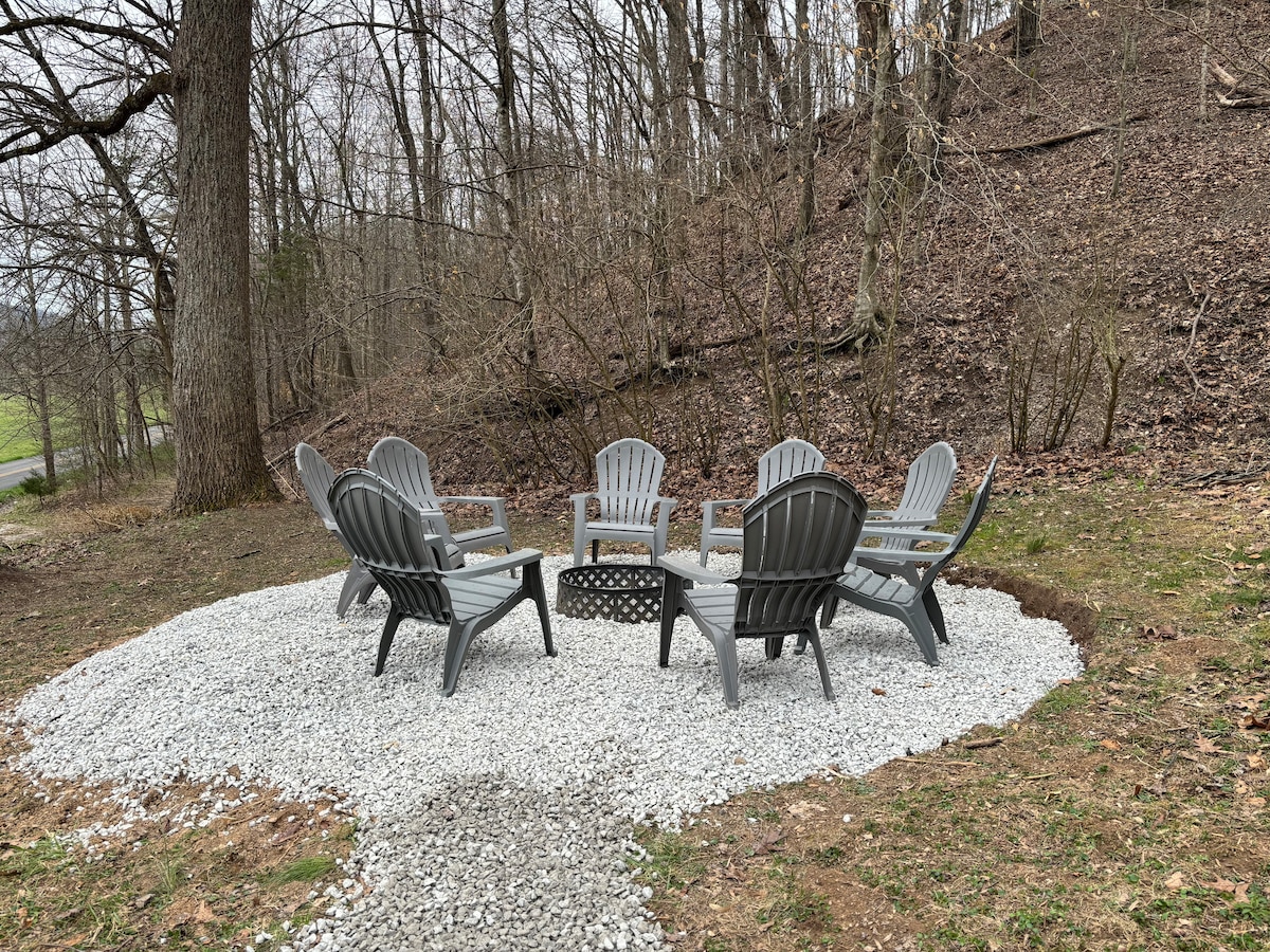 A gravel area is surrounded by a circle of eight gray Adirondack chairs, providing a cozy gathering space. A fire pit sits centrally among the chairs, with a backdrop of bare trees and a hill, creating an inviting spot for outdoor relaxation.