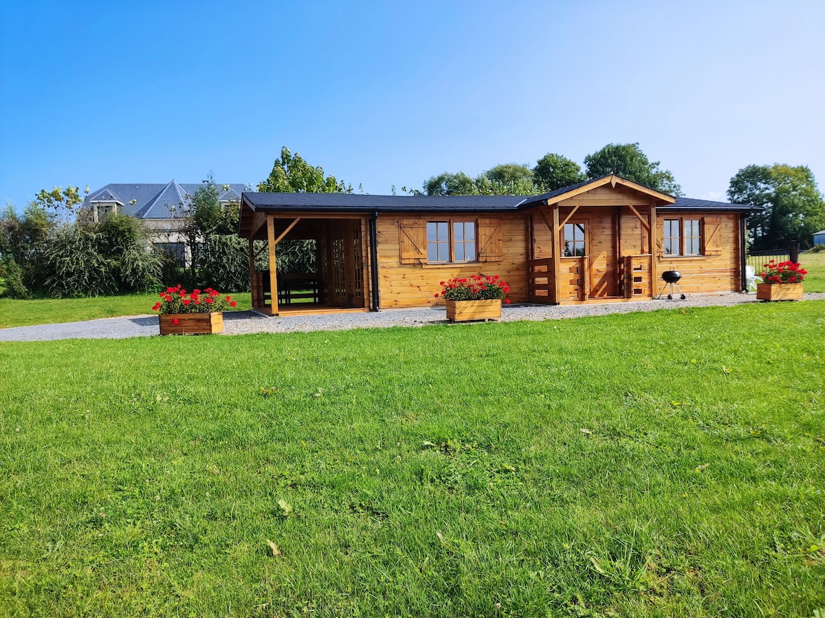 A wooden chalet is set against a clear blue sky, surrounded by green grass. Flower planters line the entrance, adding a touch of color. The structure features a covered terrace, large windows, and a welcoming facade, emphasizing a connection with the outdoor environment.