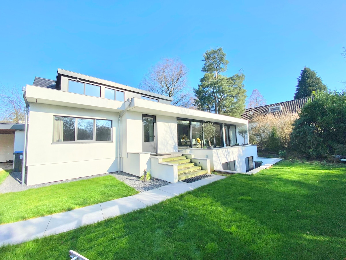 A modern house is set within a lush green yard, featuring large glass windows that open up to the outdoor space. Thoughtfully designed steps lead to the entrance, and surrounding trees provide a natural backdrop under a clear blue sky.