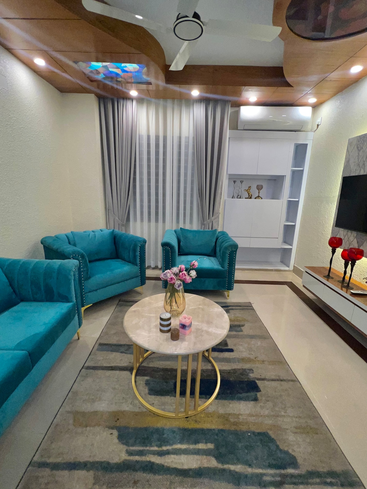 A cozy seating area is presented with two teal armchairs positioned around a round coffee table. A vase of flowers rests on the table, set against a neutral color palette. A patterned rug lies beneath, while soft light from the ceiling illuminates the space.