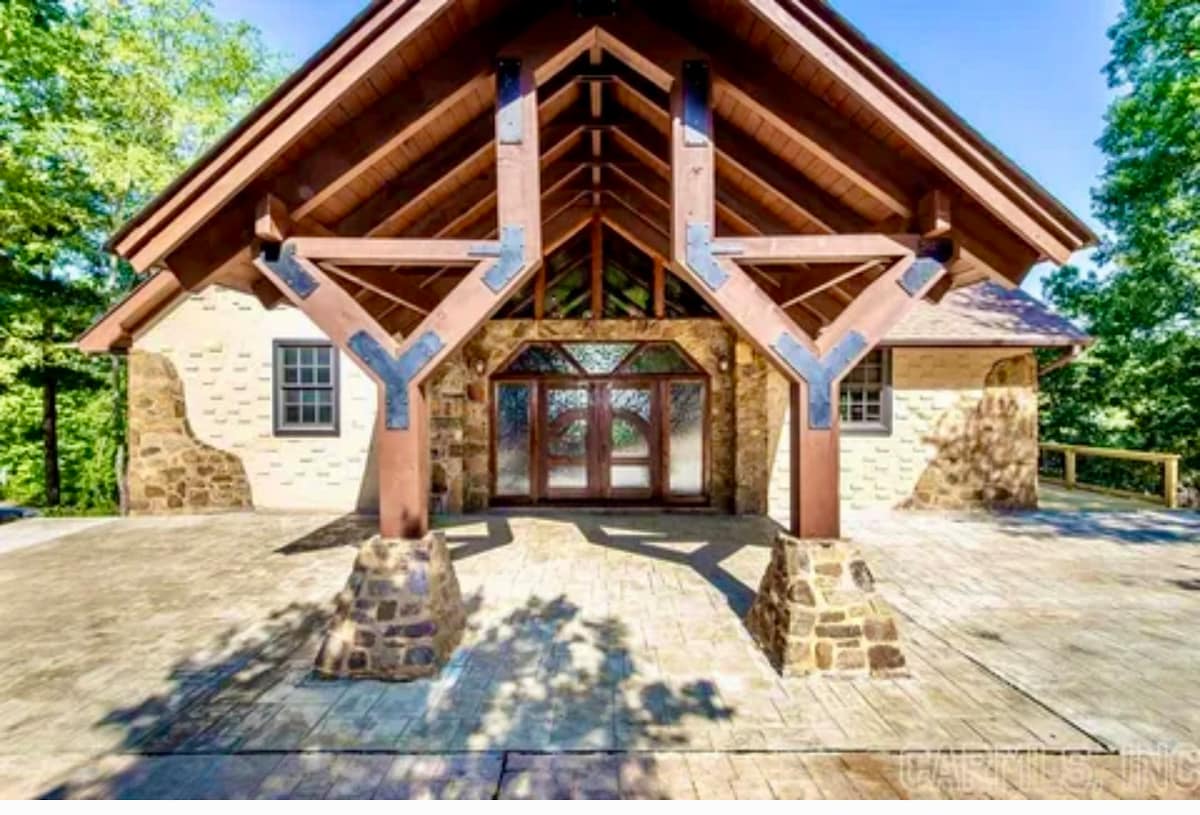 The exterior features a spacious porch supported by distinctive wooden beams and stone pillars. Large double doors invite entry, surrounded by expansive glass windows that allow natural light to fill the space. A scenic backdrop of greenery is visible in the background.