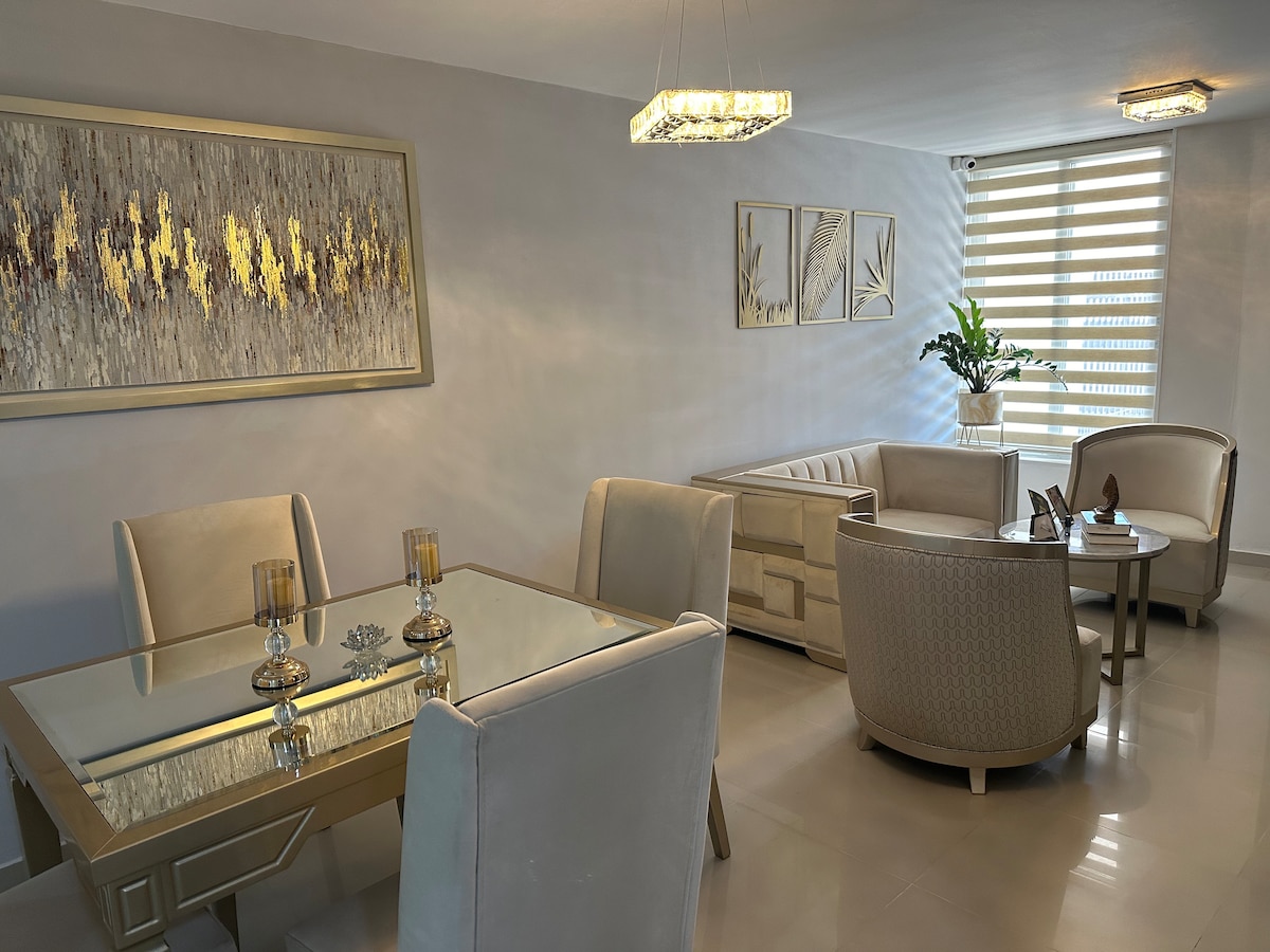 A modern living area is displayed with a glass dining table and elegant chairs. Soft lighting highlights a textured wall art piece, while large windows are partially covered by light fabric shades. Potted greenery adds a touch of nature to the inviting space.