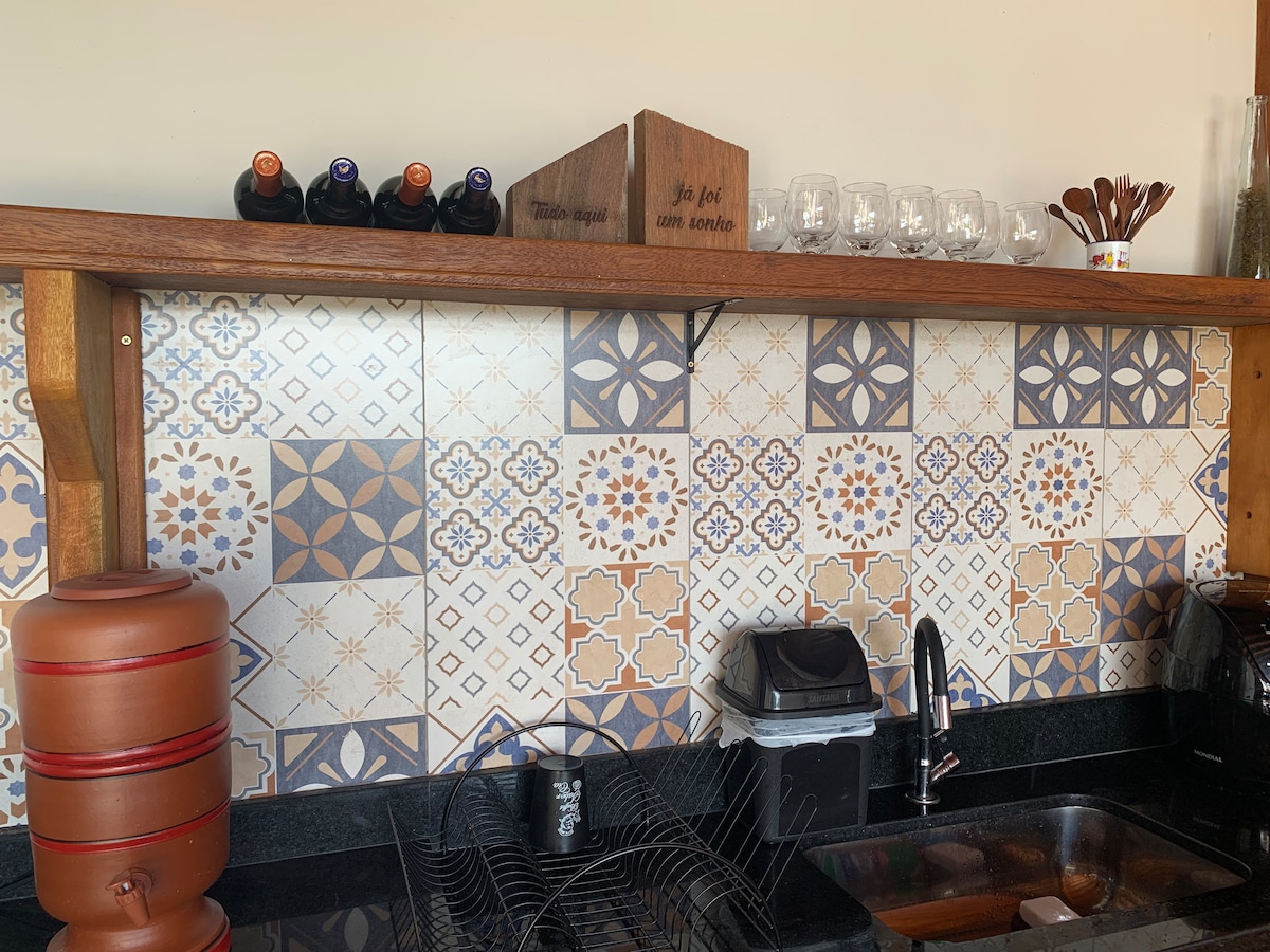 A well-organized kitchen features colorful patterned tiles on the wall, complemented by wooden shelving holding bottles and glassware. A dark countertop accommodates essential kitchen appliances, including a water dispenser and a dish rack arranged neatly.