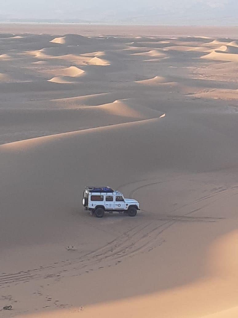 A rugged vehicle is positioned on sandy terrain, with gentle dunes stretching across the landscape in the background. The soft contours of the sand are highlighted by natural shadows, creating a serene atmosphere. The vastness of the desert is clearly visible, extending towards distant horizons.