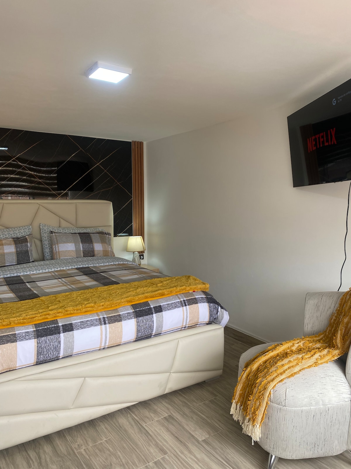 A contemporary bedroom features a neatly made bed with patterned bedding and a warm throw at the foot. A flat-screen TV is mounted on the wall, while a modern chair is positioned nearby, providing a cozy spot for relaxation.