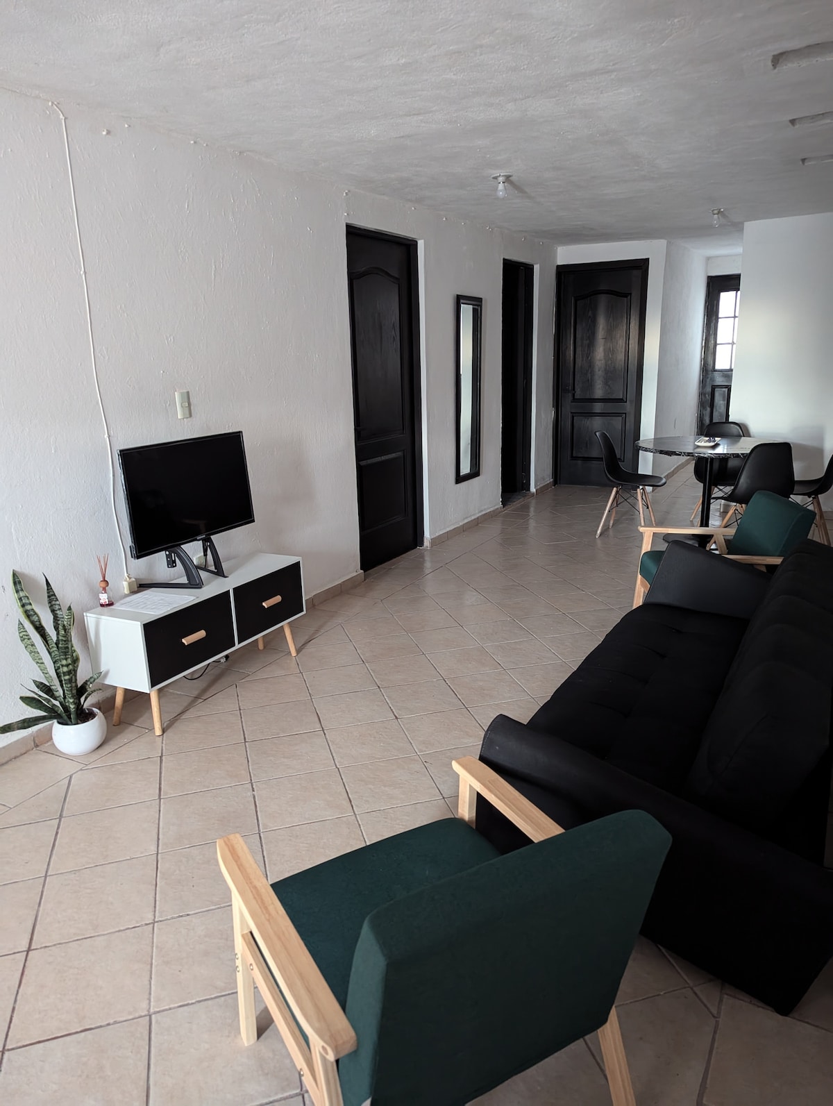 A living area features a black sofa, two green chairs, and a television on a white cabinet. Natural light illuminates the space through a nearby window, and a plant adds a touch of greenery. A dining table is visible in the background, enhancing the open layout.