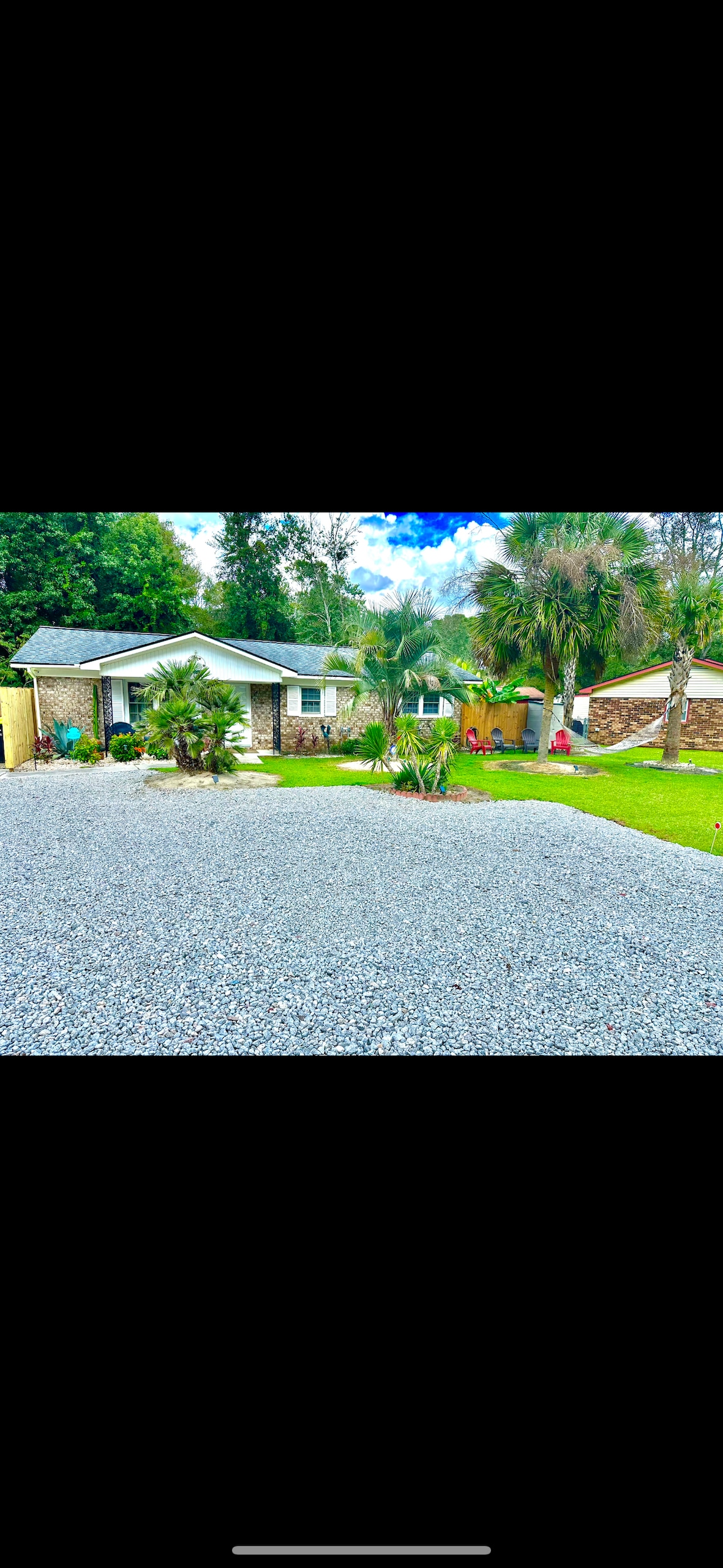 A welcoming exterior of the home is showcased, featuring a gravel driveway leading to a well-maintained lawn with various shrubs and greenery. The house is framed by palm trees and other plants, creating a relaxed atmosphere in a central location.