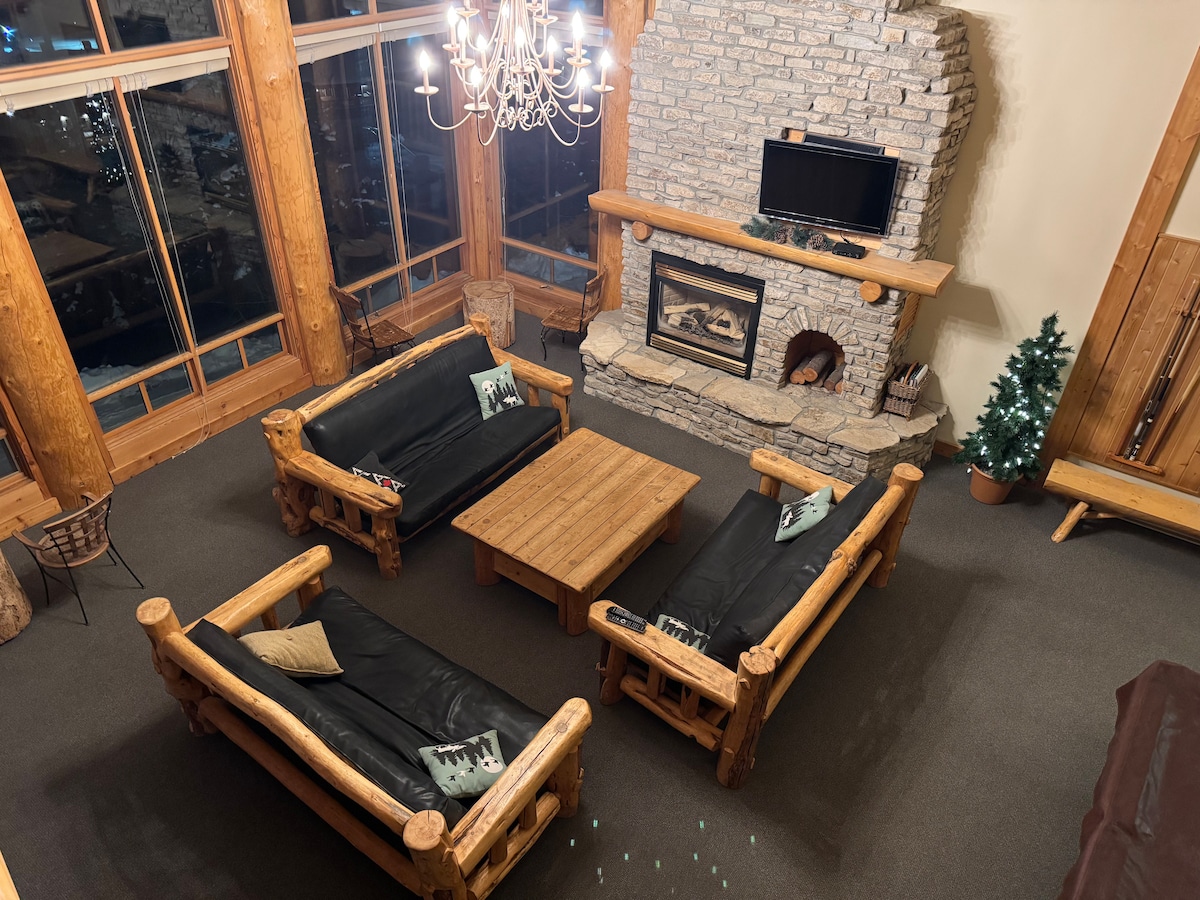 A spacious great room features rustic log furniture, including four black leather couches surrounding a wooden coffee table. A stone fireplace is centered against the wall, and large windows allow natural light to fill the space, showcasing views of the surrounding landscape.