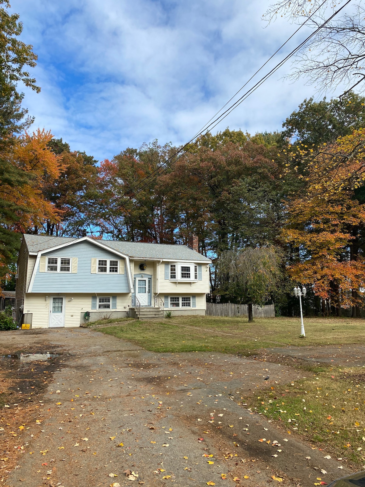 A two-story house is set among colorful autumn trees, with a spacious front yard covered in fallen leaves. The exterior features light blue siding and a welcoming entryway. Above, a bright sky filled with clouds adds a serene backdrop to the home.