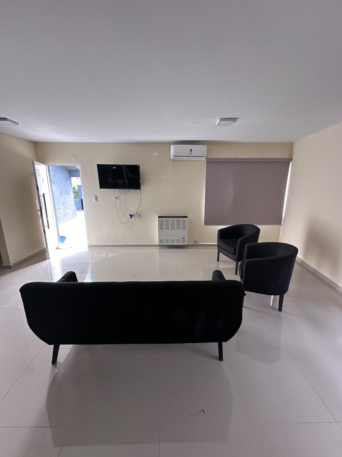 A modern living area features a black sofa and two black armchairs, positioned around a wall-mounted TV. Natural light enters through a door on the left, and a window is covered with a light shade. Light-colored walls and glossy floors enhance the spacious feel.
