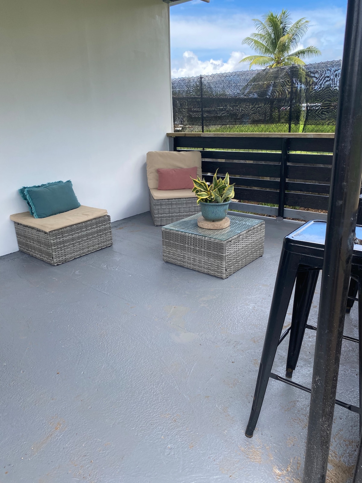 A small outdoor seating area features two cushioned chairs beside a glass-top table. A potted plant is centered on the table. The space is surrounded by a black wooden fence, with a view of greenery in the background.