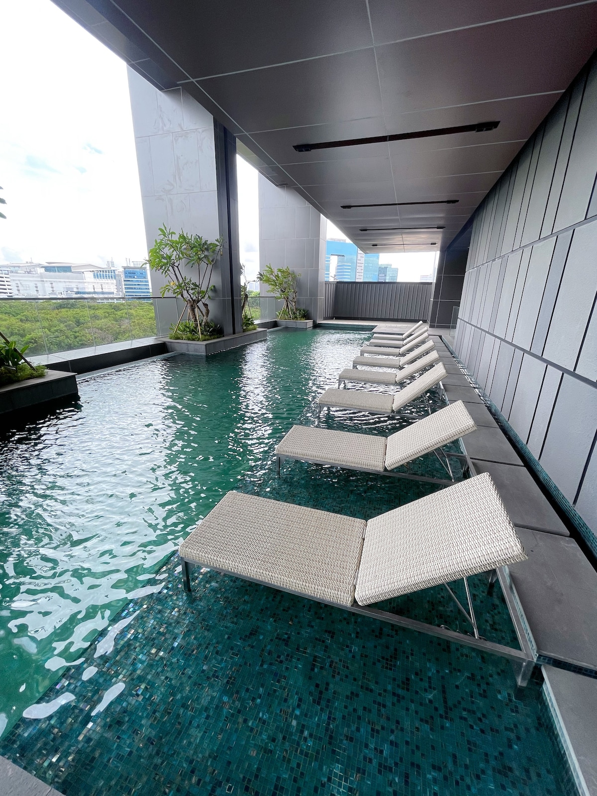 A sleek swimming pool is seen on a balcony, surrounded by modern grey paneling. Several lounge chairs are positioned along the water's edge, inviting relaxation. Planters with greenery add a touch of nature, with city views visible beyond the pool area.