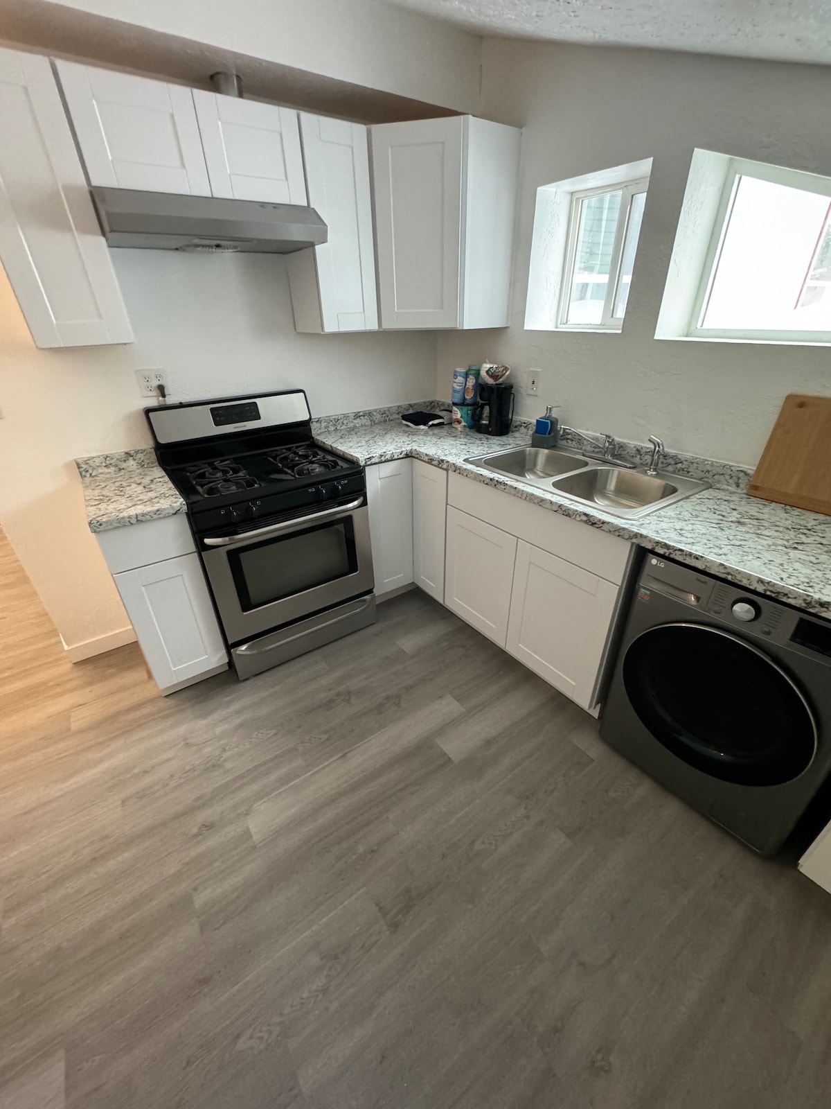 A modern kitchen is presented, featuring white cabinets and sleek granite countertops. Stainless steel appliances include a gas stove and a washer-dryer combo, positioned beside a spacious sink. Natural light streams through two nearby windows, enhancing the bright, functional space.