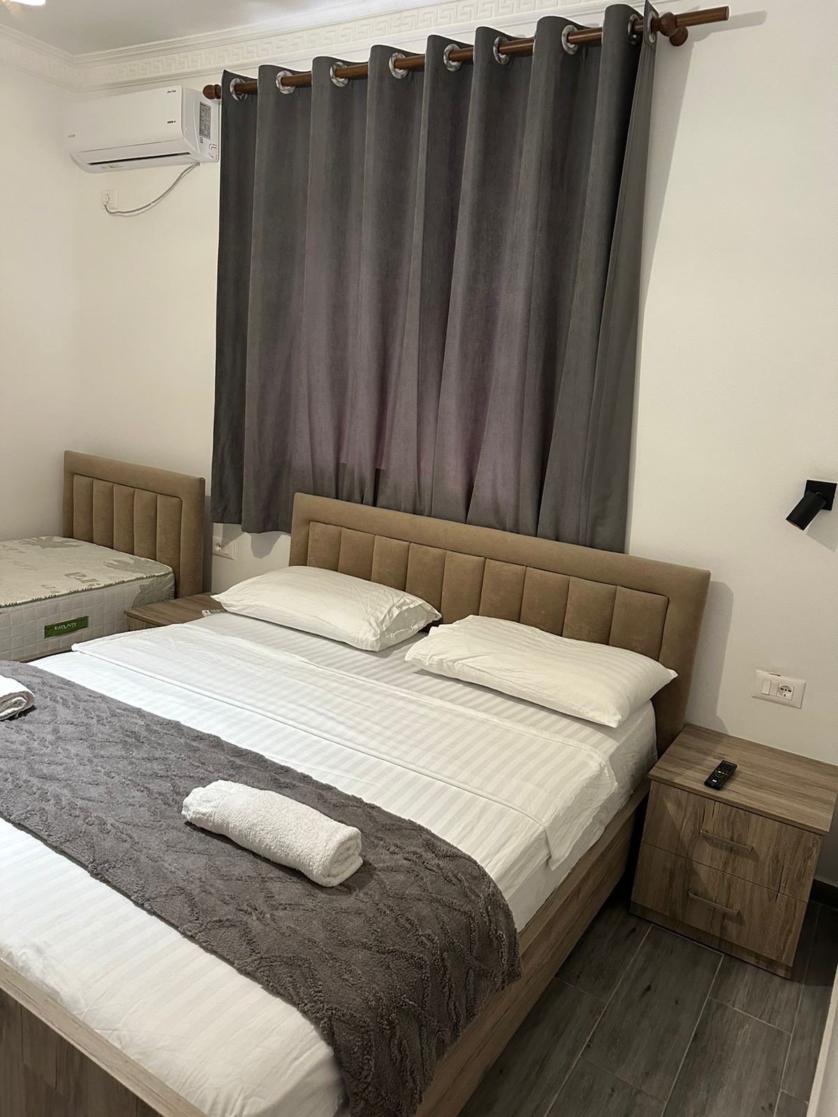 A sleeping area features a spacious bed with neutral-colored linens and a textured throw. Two padded headboards complement the arrangement. A bedside table holds a small lamp. Dark curtains filter light from a window, enhancing the tranquil feel of the room.