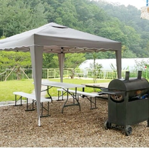 A shaded outdoor seating area is provided by a large gray canopy. A series of long tables are set beneath it, accompanied by a barbecue grill positioned nearby. Lush greenery and distant structures can be seen in the background.