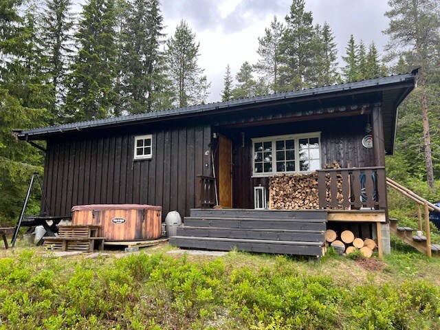 A rustic cabin is nestled among trees, featuring a dark wooden exterior and a welcoming entrance. A hot tub is located on the porch, beside neatly stacked wood. The cabin is surrounded by a natural landscape with greenery and gentle slopes.