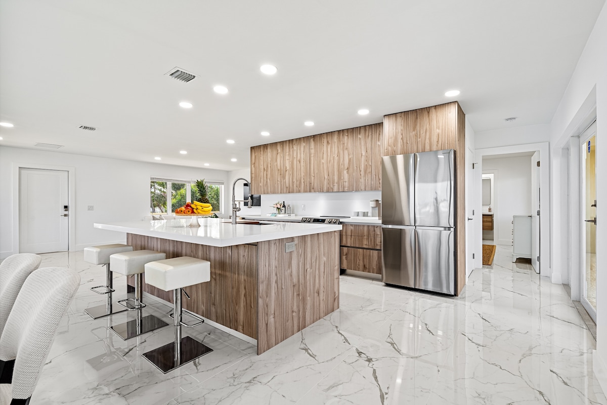 A modern kitchen is displayed with a large island featuring seating and a sleek countertop. Stainless steel appliances are integrated with wooden cabinetry. Bright lighting highlights the spacious, open layout, complemented by polished marble flooring. Windows offer natural light and views of the exterior.