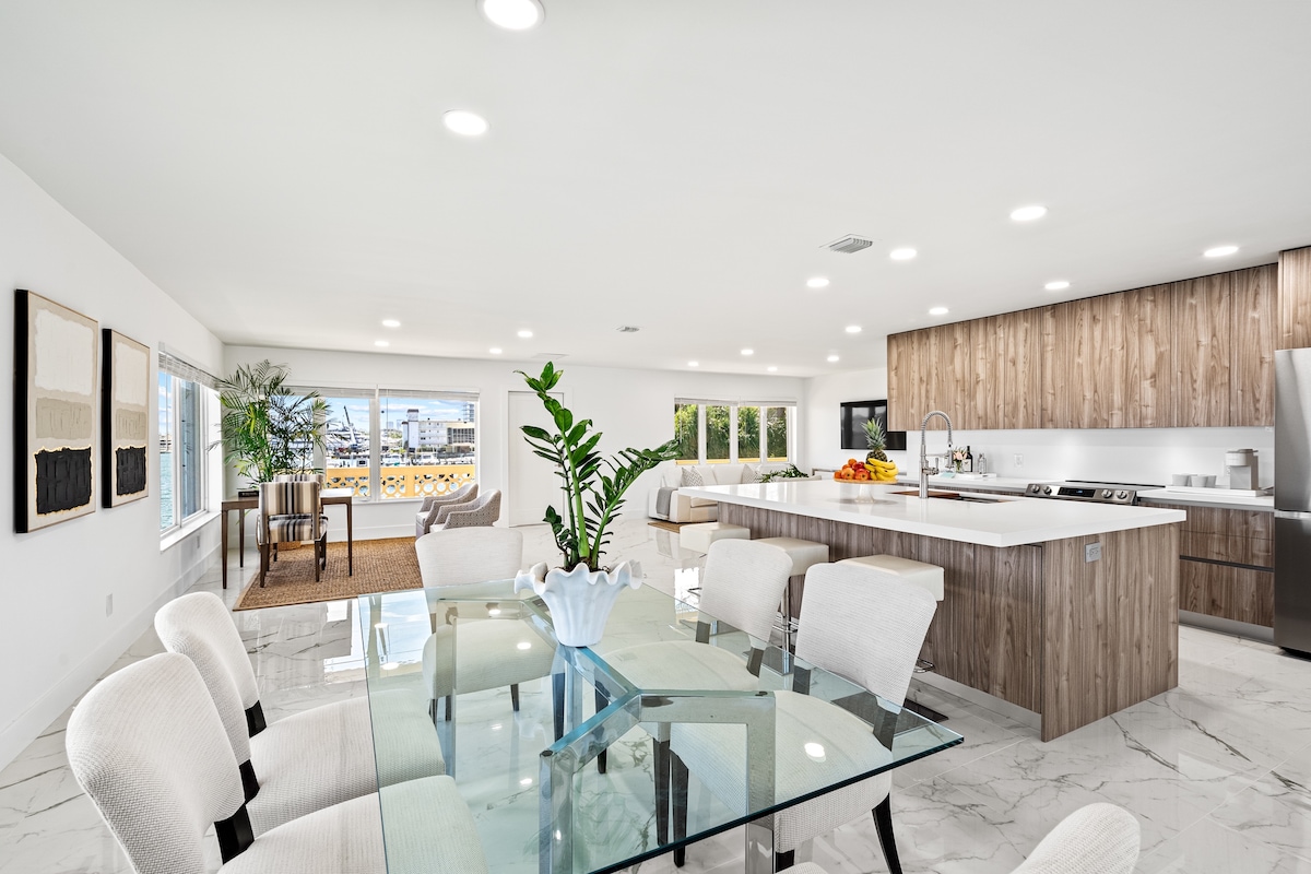 An open-concept living area is featured, highlighting a modern kitchen with high-end appliances and a central island. A dining table with light-colored chairs is situated nearby, complemented by large windows that allow natural light to fill the space and offer views of the surroundings.