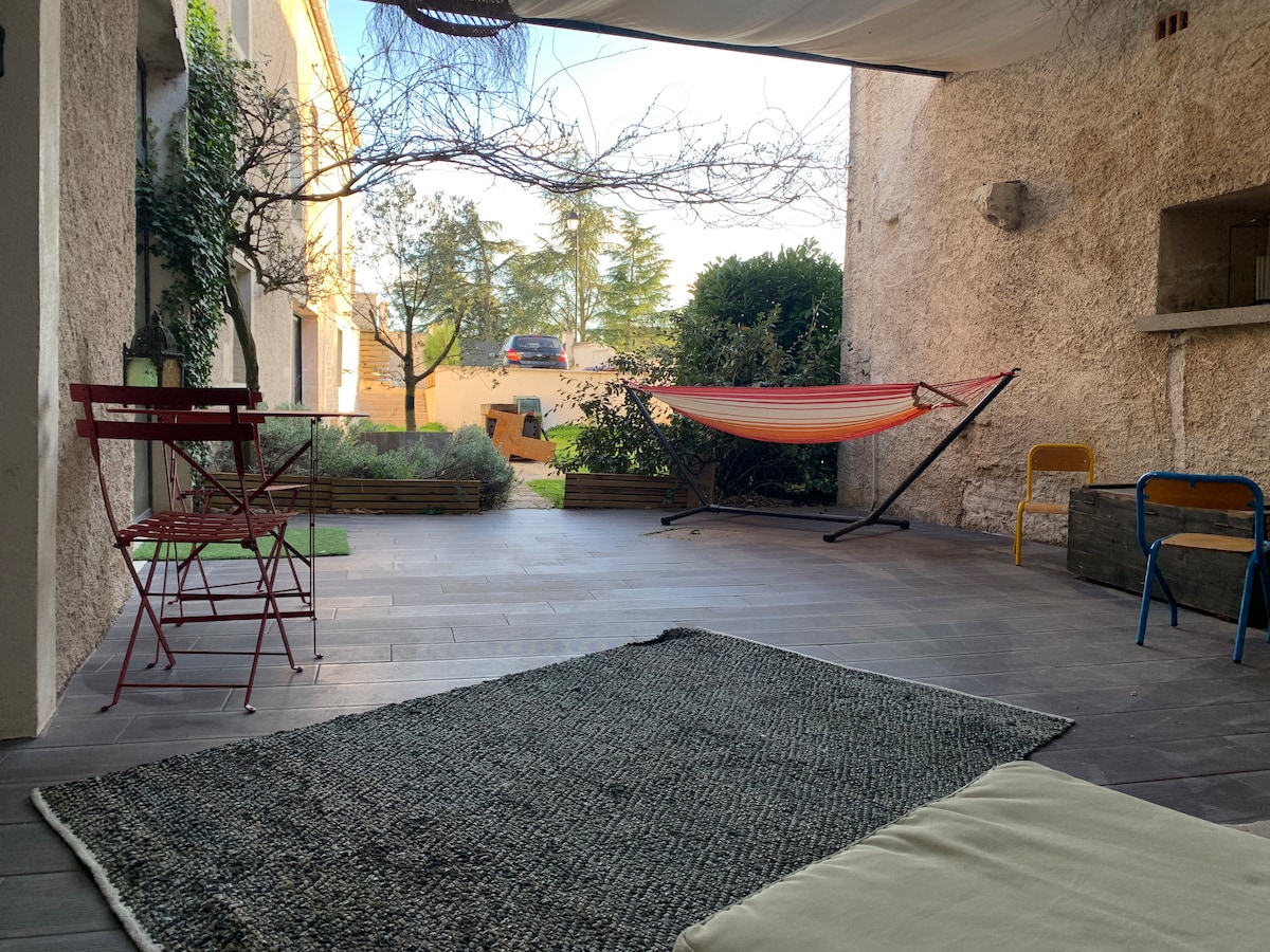 A covered outdoor space features a hammock, a small red table with two chairs, and a neutral-colored rug. Greenery and trees are visible in the background, enhancing a peaceful atmosphere. Sunlight filters through, creating a warm and inviting environment.