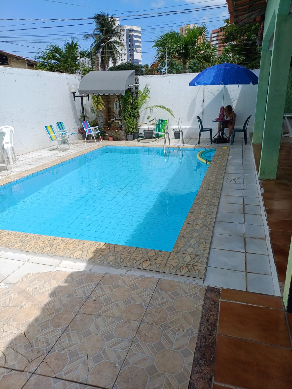 A serene outdoor area features a swimming pool with clear blue water, surrounded by tiled flooring. A seating area with two chairs and a table is positioned under an umbrella, while colorful lounge chairs are arranged nearby, enhancing the space for relaxation.