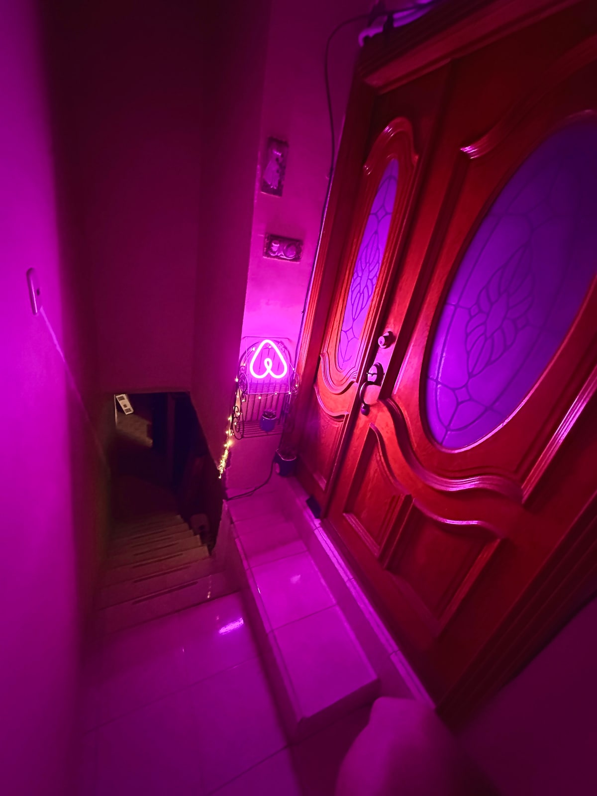 The entryway features a wooden door with decorative glass panels, illuminated by soft purple lights. A small staircase leads downward, with the walls painted in a muted tone, creating a welcoming atmosphere. Neon heart-shaped signage adds a modern touch to the space.