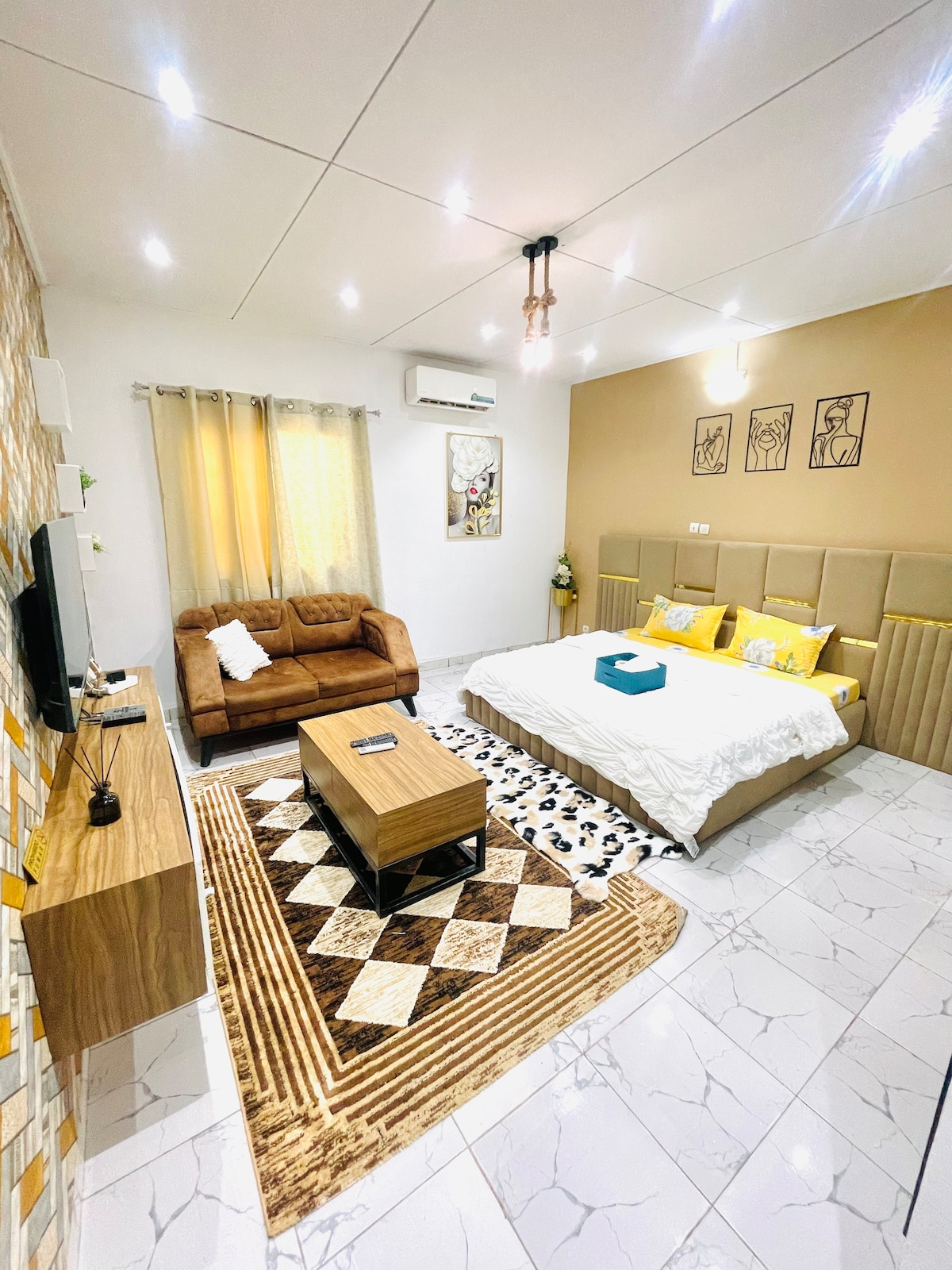 A modern living space features a spacious bed with soft bedding and decorative pillows. A comfortable brown sofa sits adjacent to a wooden coffee table. Warm lighting enhances the cozy atmosphere, while large windows are dressed with light curtains.