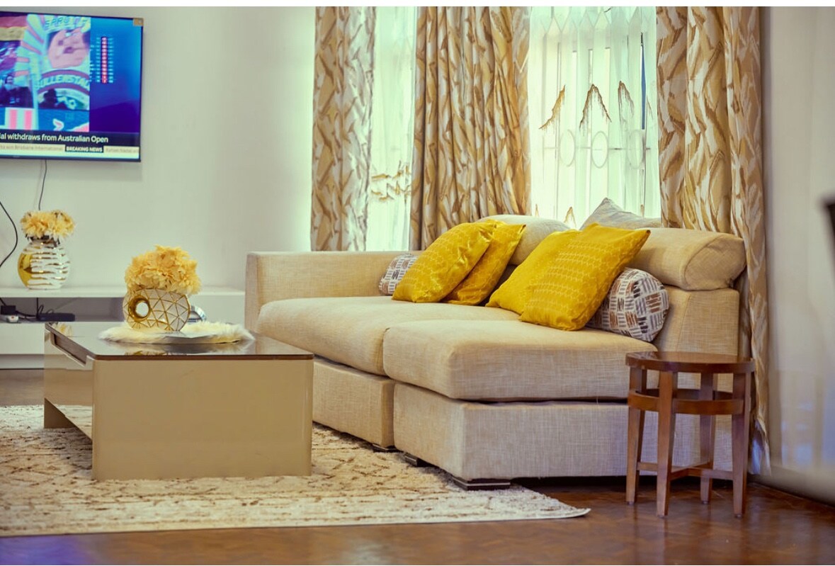 A neutral-toned sofa invites relaxation, adorned with yellow accent pillows. A decorative coffee table with a geometric centerpiece is positioned in front, complemented by a small side table nearby. Soft, flowing curtains frame the window, and a television is mounted on the wall.