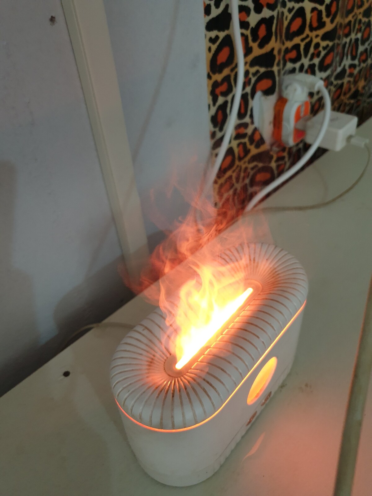 A modern mini heater is visible on a surface, emitting a warm orange glow. The device features a sleek design, with swirling patterns in the background adding a touch of texture to the space. Nearby, multiple electrical outlets and cords are present.