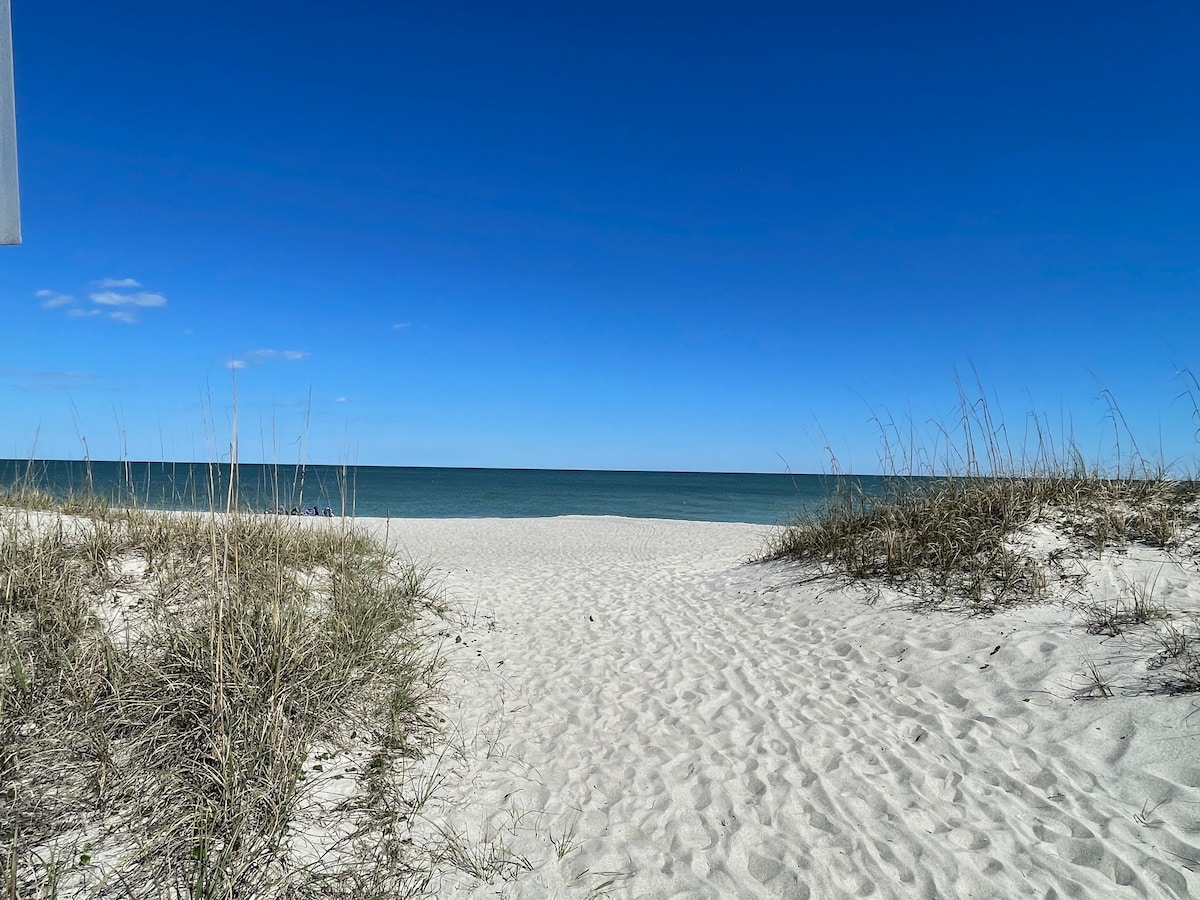 A sandy path leads from a grassy area to the tranquil beach, framed by soft dunes. The horizon showcases a clear blue sky, seamlessly merging with the calm ocean. Sunlight glistens on the water, inviting relaxation and leisure.