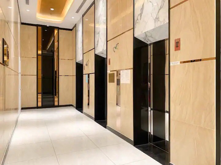 A modern elevator lobby features polished marble accents and sleek reflective surfaces. Soft lighting highlights the clean lines of the space, while a series of elevators is positioned along one side. The white tiled floor contrasts with the darker elements, creating a sophisticated atmosphere.
