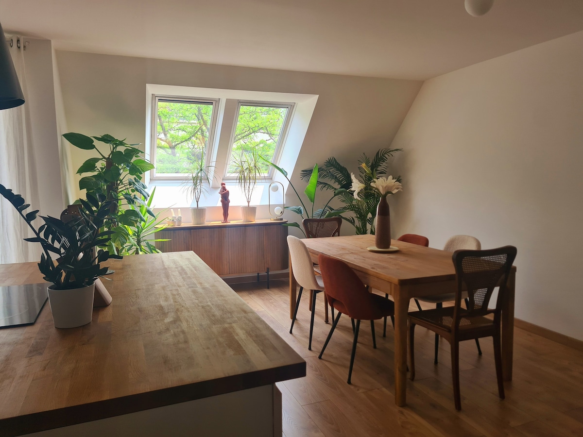 A bright living area features large angled windows that invite natural light and showcase lush greenery outside. A wooden dining table is surrounded by various chairs, while decorative plants are positioned throughout the space. A sideboard adds additional storage and character to the room.
