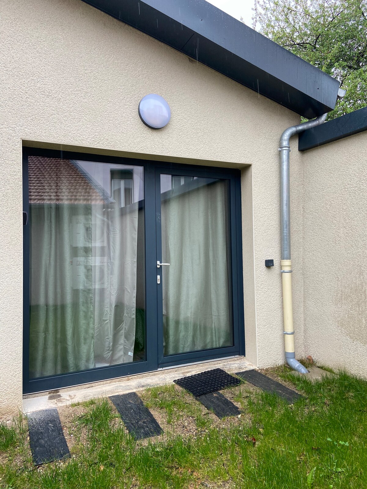 A sleek entrance is shown, featuring a set of glass double doors framed in dark materials. Semi-transparent curtains are visible, allowing glimpses of greenery outside. A simple welcome mat is positioned in front, and the surrounding area is bordered by a patch of grass and stone pathway.