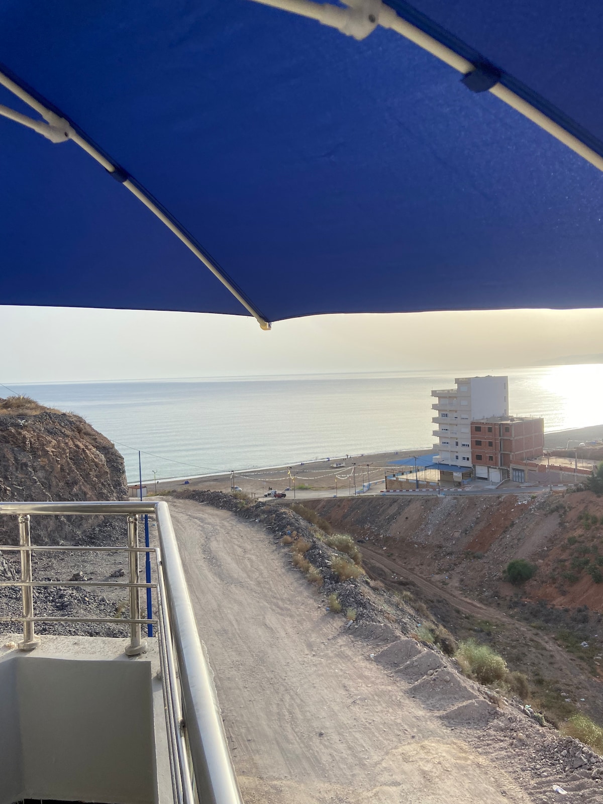 A panoramic view of the coastline is framed by a blue umbrella. The gently sloping shoreline and calm sea are visible in the distance, with a distant building positioned along the beach. A gravel path leads towards the water, surrounded by rocky terrain.