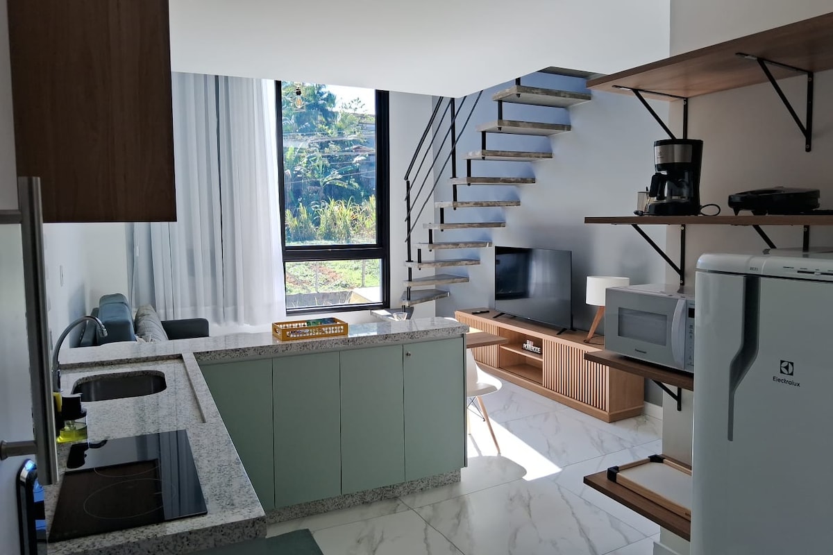 A modern kitchen area features sleek cabinetry and a granite countertop, displaying a built-in stovetop and a microwave. A cozy living space includes a sofa and a TV stand, with large windows allowing natural light to illuminate the room.