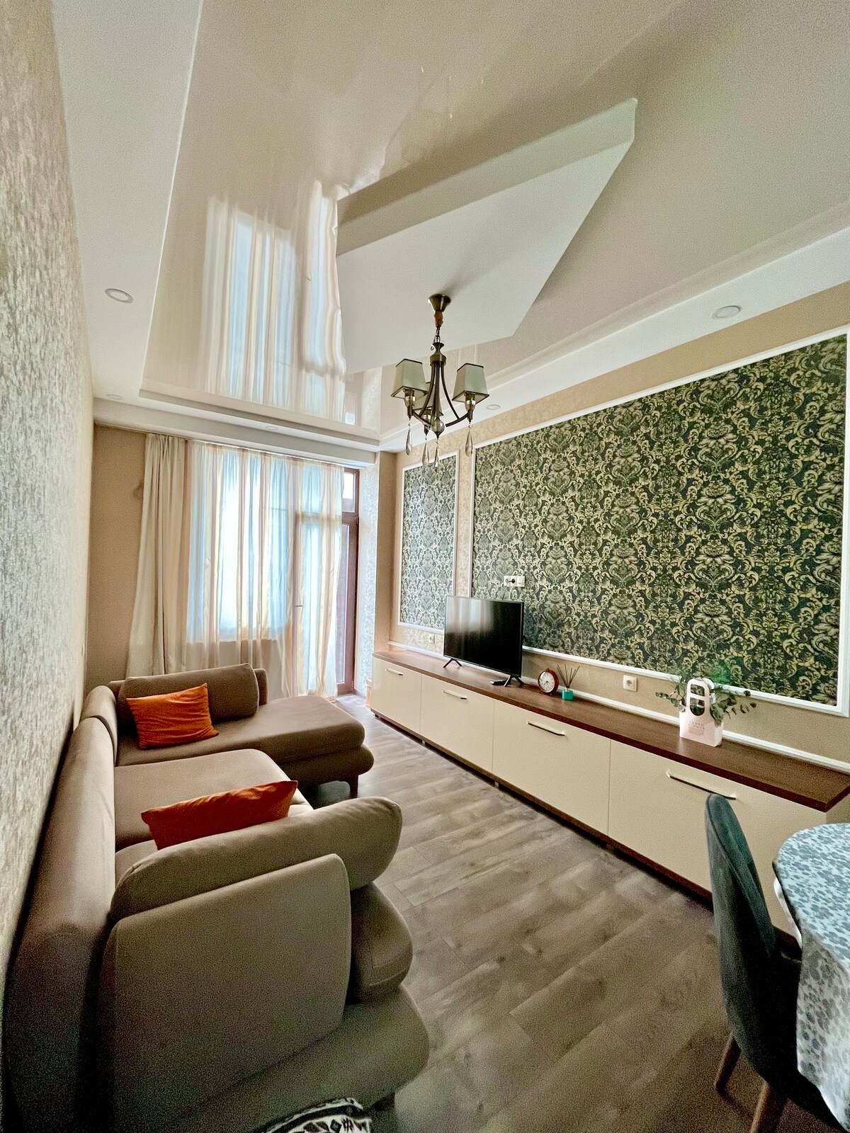 A bright living area features a comfortable sofa and decorative cushions. Elegant wallpaper and a modern chandelier enhance the space, while a television is mounted on a console beneath decorative wall panels. Natural light filters in through large windows with sheer curtains.