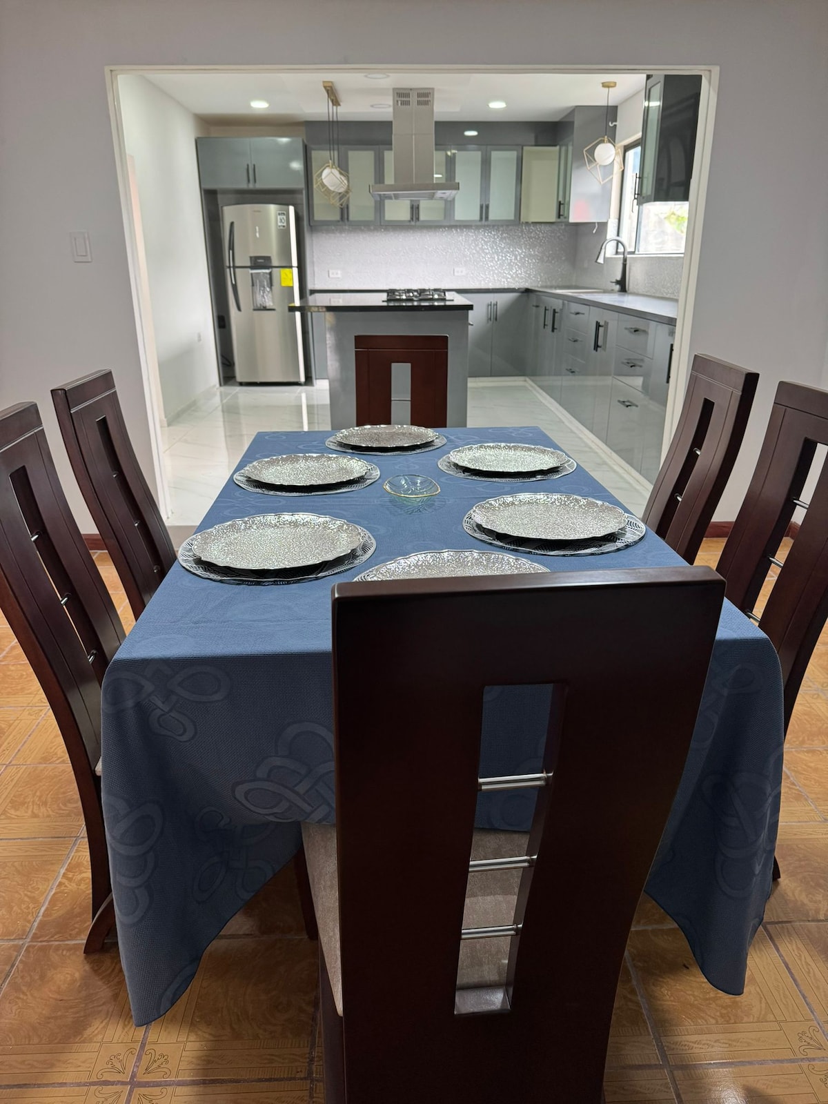 A dining area features a wooden table set with a blue tablecloth and six decorative plates. Modern kitchen spaces are visible in the background, showcasing sleek cabinets and stainless steel appliances. Natural light enters through a window, creating a bright and inviting environment.